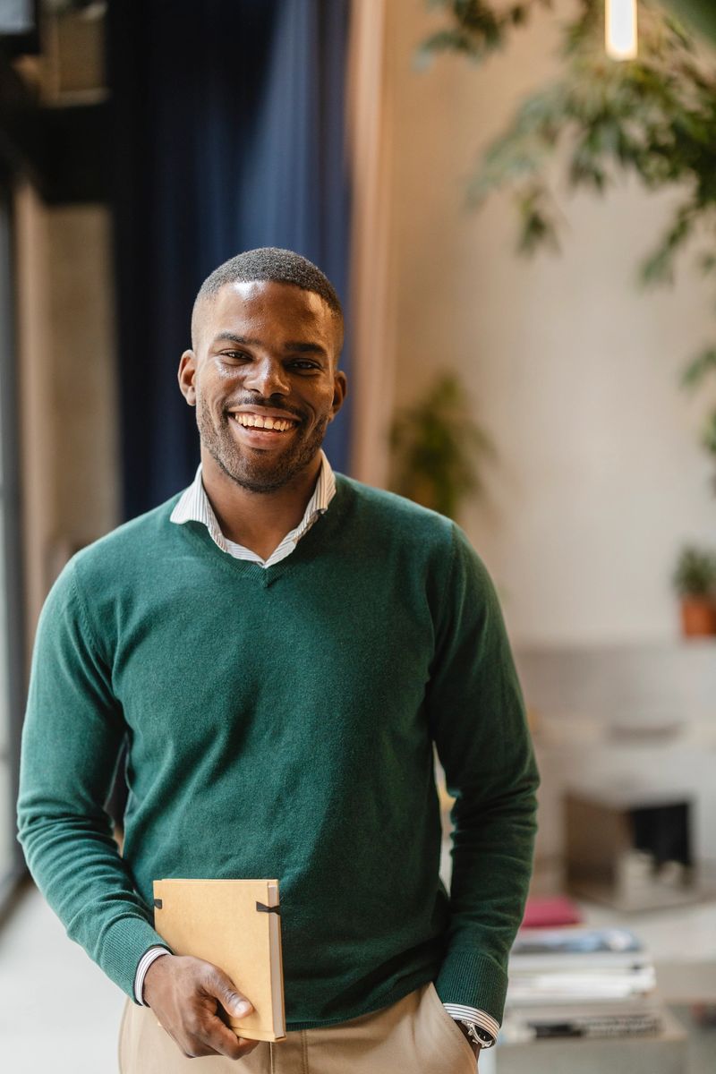 Young, happy african american educator stands in a brightly lit, modern classroom, holding a notebook and smiling confidently, ready to inspire his students