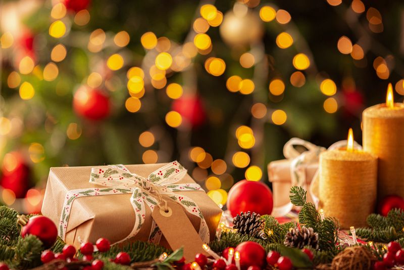 Christmas gift box with a blank tag wrapped with brown craft paper on a cozy table with Christmas decore candles and illuminated tree at background. Gift or surprise, New Year's Eve. Copy space