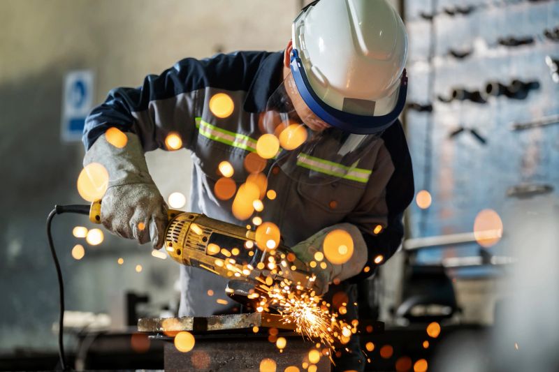 Worker in protective suit operates heavy-duty grinding equipment in a factory setting. Flying sparks illuminate the workshop, showcasing strength, precision, and occupational safety in a demanding metalworking environment.