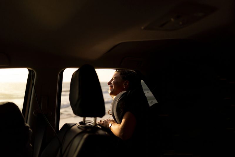 Carefree young woman leaning out of car window, enjoying a beautiful sunset at the beach