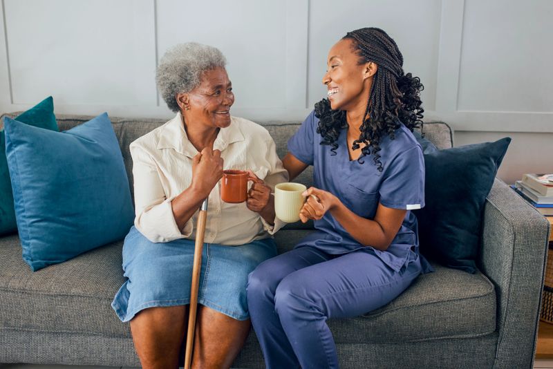 Front view of happy senior woman and young nurse smiling and having conversation holding mugs on sofa at home