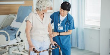 Nurse helping an older woman with a Zimmer frame and hospital bed in the background.