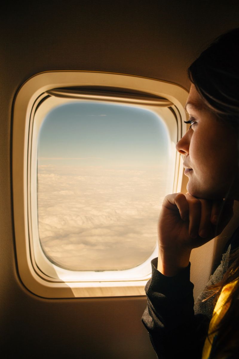 A thoughtful woman gazes out of an airplane window, enjoying the serene view of clouds and the blue sky.