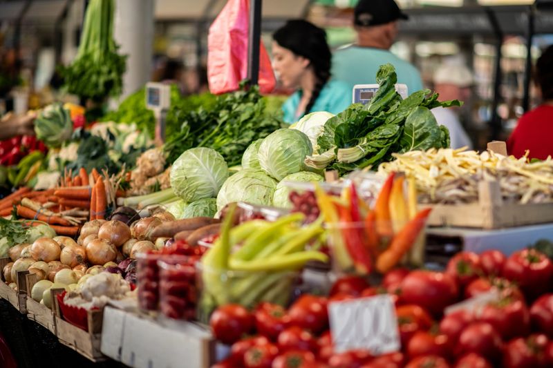 Market stall overflowing with fresh vegetables in every color—tomatoes, peppers, leafy greens, and more. A glimpse of a woman working behind the scale adds to the authentic market atmosphere.