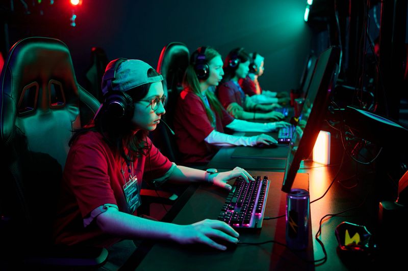 Group of multiethnic teenage girls wearing headsets competing in esports tournament, sitting in row at gaming computers, focused on playing video game together as team in dark room