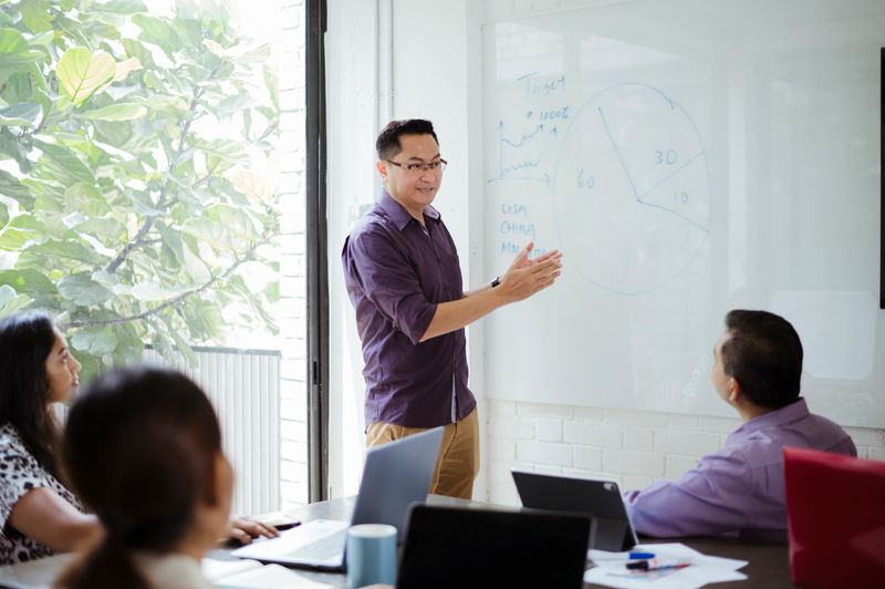 A mature businessman conducts a team meeting, using charts and graphs to explain performance targets and company direction. The scene highlights knowledge sharing, team leadership, and collaboration.