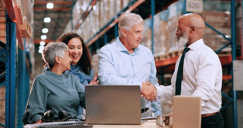 Business people, laptop and handshake in warehouse for logistics, meeting for supply chain collaboration. Import team, tech or shaking hands in storage for export deal, service agreement or welcome