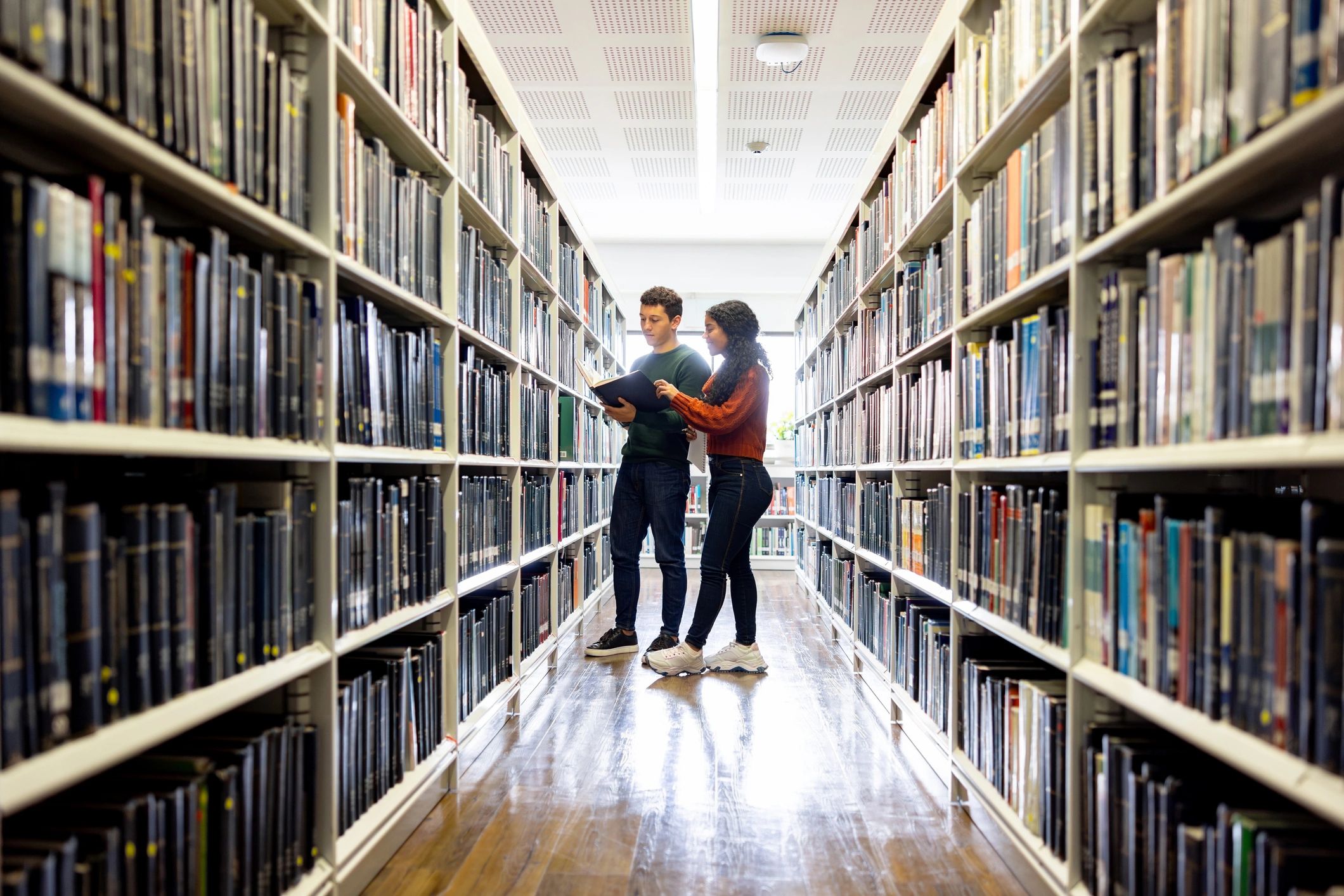 Two young people reading a book in a library aisle.