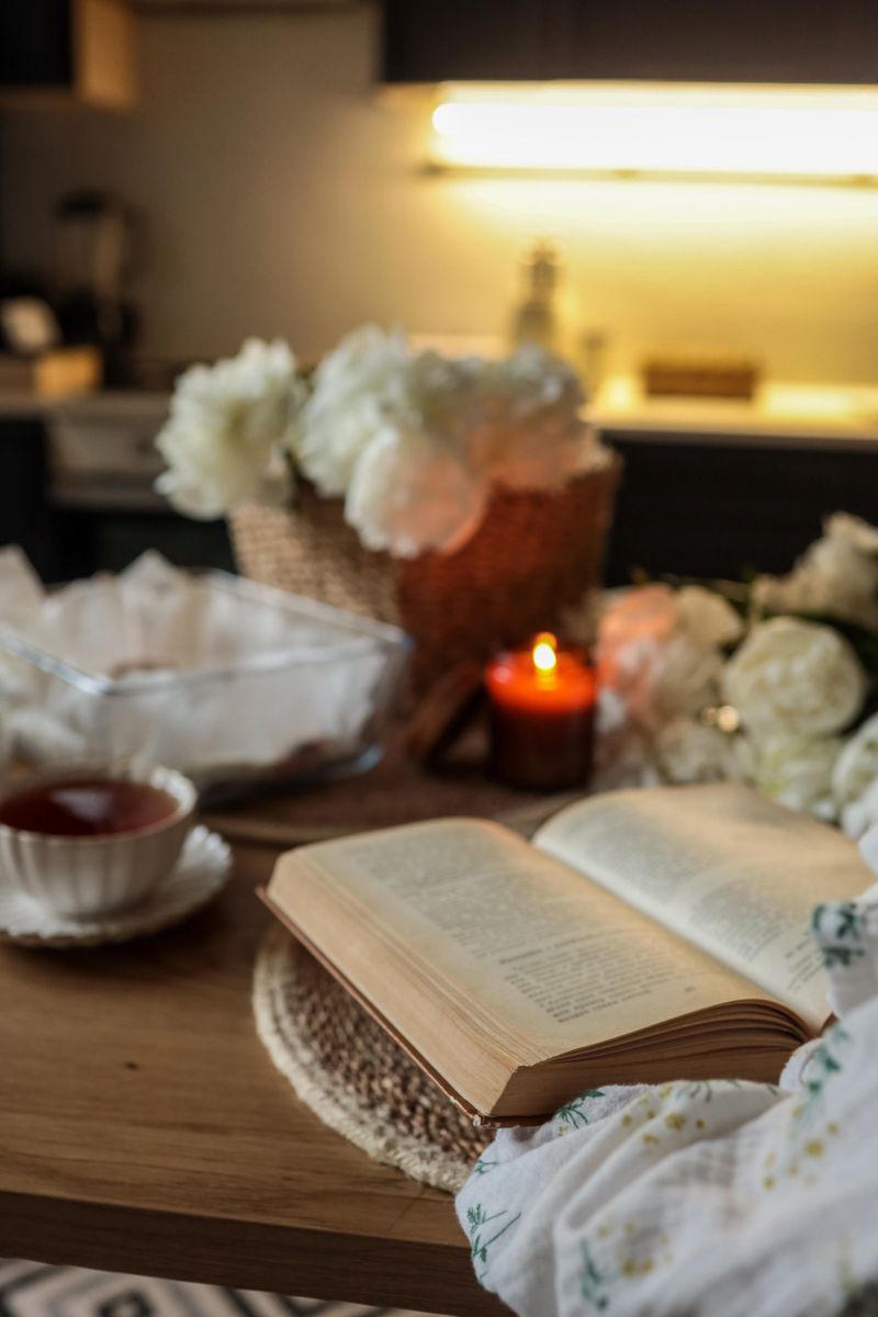 Open book on a wooden table beside a cup of tea, candle, and peonies in a cozy kitchen setting. Soft lighting and woven textures create a warm, peaceful reading atmosphere.
