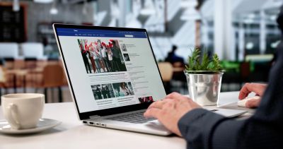 Person browsing news on a laptop with a plant and coffee on the table.