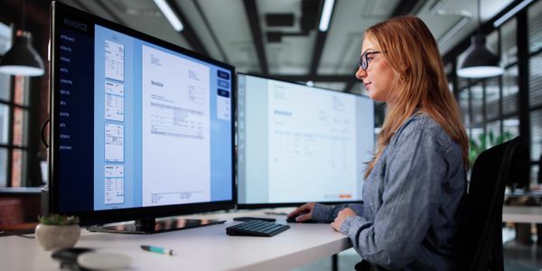 Woman reviewing invoices on dual monitors in a modern office.