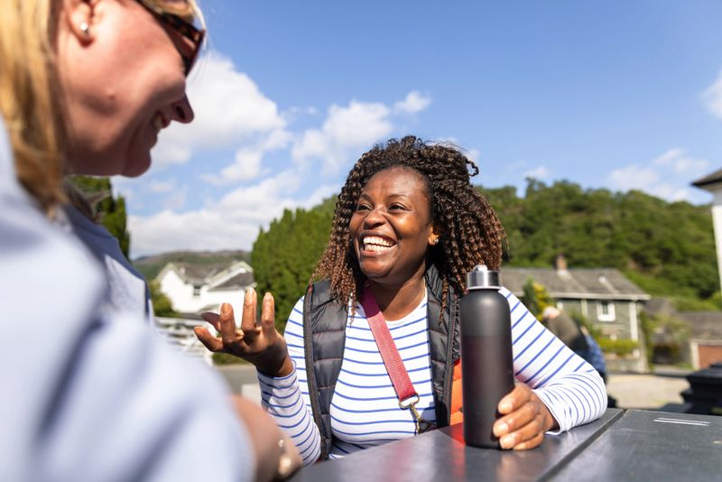A medium over-the-shoulder shot of two mature female friends sitting down outside a cafe on a picnic bench. They are both looking at each other and smiling while they engage in a conversation. One woman is holding a black water bottle. Videos are available similar to this scenario.