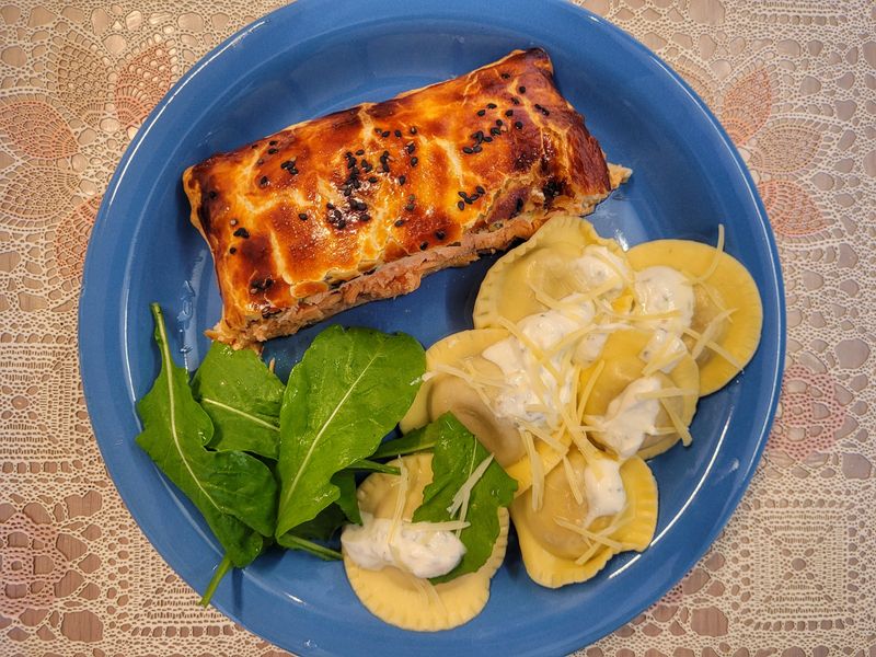 A plated dish featuring Salmon Wellington made with golden puff pastry, filled with fresh salmon, spinach, artichoke hearts, and artisanal cream cheese. Served with Uruguayan-style sorrentinos stuffed with mushrooms, topped with a creamy four-cheese sauce and finished with shaved parmesan. The salad includes fresh organic arugula leaves. A sophisticated, well-balanced, and visually appealing meal