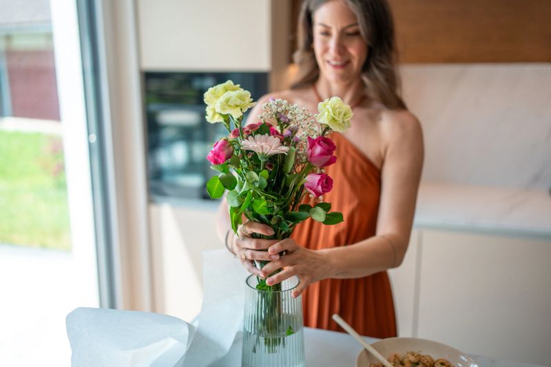 A mid adult Caucasian woman in an orange dress arranges a vibrant floral bouquet in a glass vase within a bright modern kitchen. Soft natural light highlights the serene and organized indoor setting.
