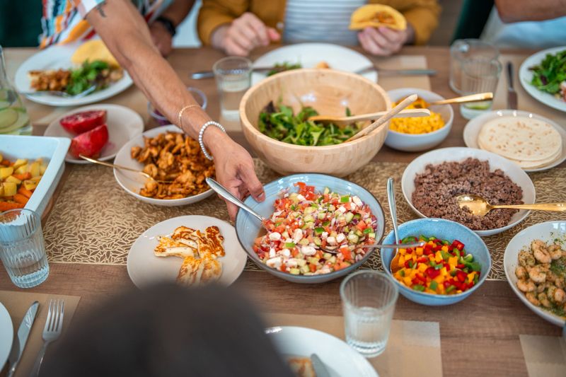 Overhead view of family-style Mexican dinner spread featuring fresh tacos, colorful salsas, black beans, and assorted toppings arranged on rustic wooden table with casual table settings.