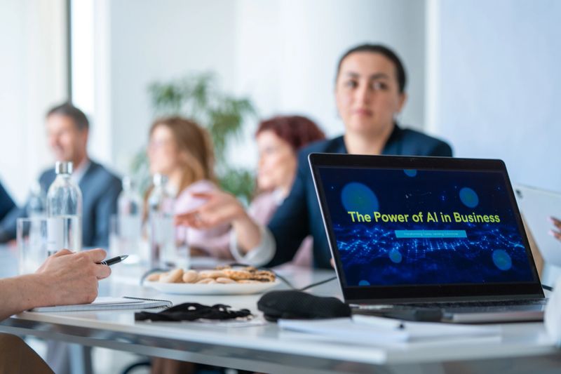 Diverse business professionals seated in a modern boardroom interact during a laptop-based AI presentation. The scene includes a sleek workspace, casual business attire, and attentive collaboration, emphasizing technology's role in business innovation.