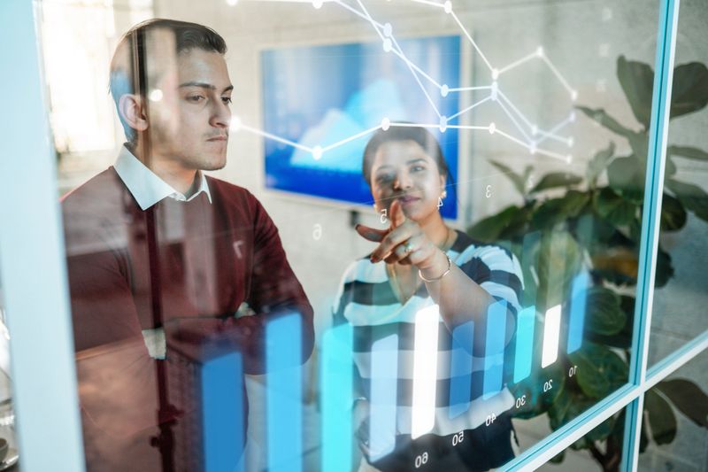 A young Hispanic male and mid-adult Indian female executives collaborate using advanced holographic data visualization tools in a sleek, modern office setting illuminated by bright, natural light.