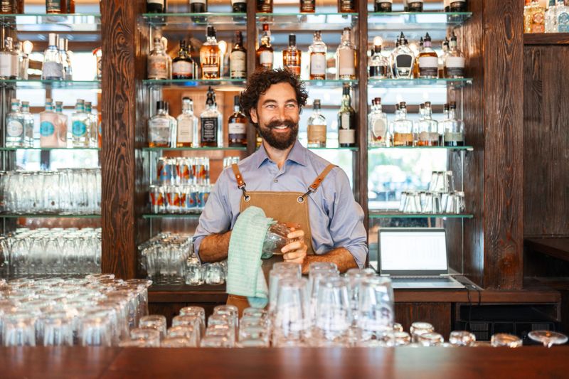 Friendly Young Bartender Smiling While Polishing Glasses