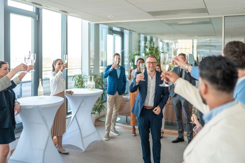 A group of adults dressed in business attire are joyfully toasting with champagne in a modern office setting. The atmosphere is festive and professional, highlighting successful business achievements and camaraderie among colleagues.