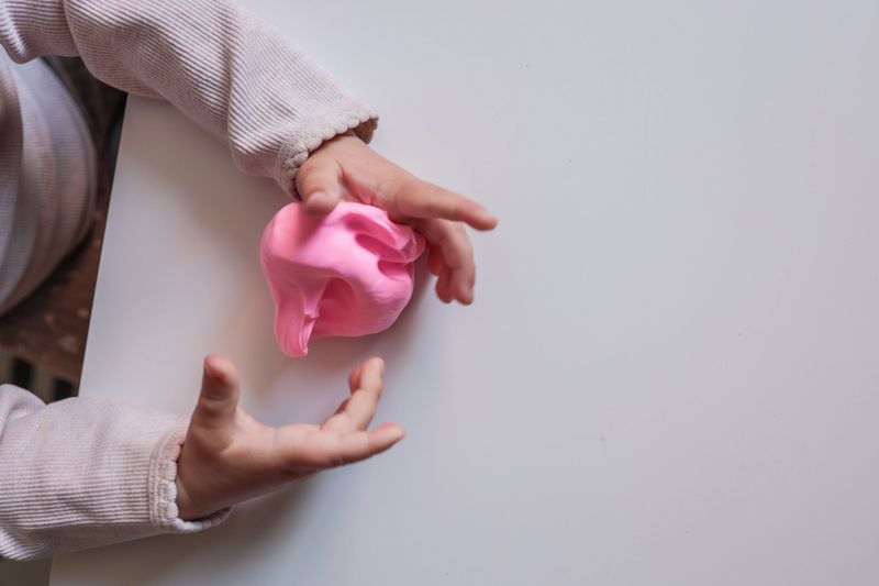 Small child squeezing soft pink modeling clay on a white table. Close-up of hands engaged in sensory and creative play, encouraging tactile exploration and motor skills.