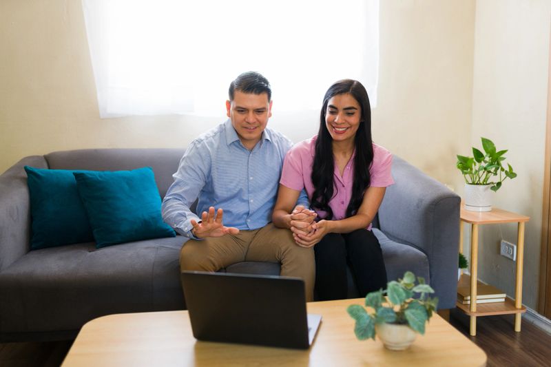 Happy latin couple holding hands and waving while having virtual therapy session with psychologist on laptop at home
