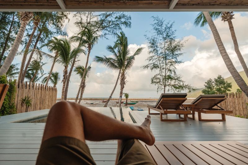 POV of a tourist relaxing in a tropical bungalow, legs crossed, taking in the view of the beach and swaying palm trees.