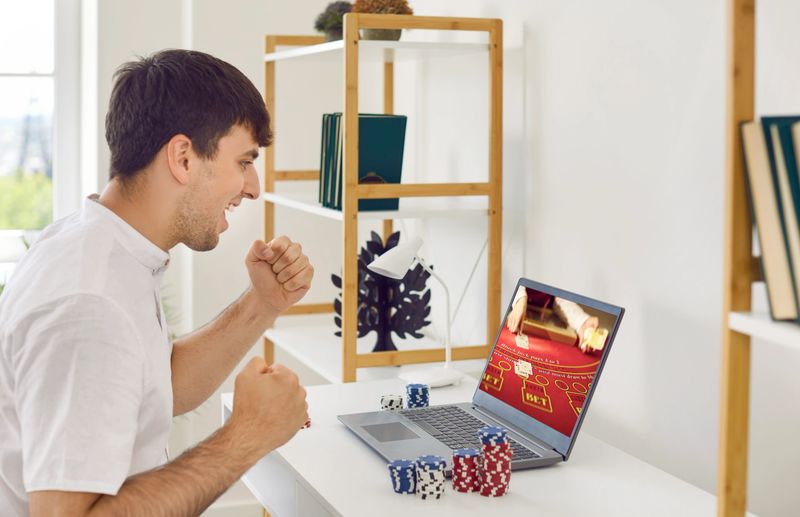 Winning online. Excited guy is amazed at big win he won playing online poker with help of laptop. Man clenches his fists with joy while sitting in front of laptop at home. Concept of online gambling.