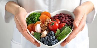 Hands holding a heart-shaped bowl with healthy fruits, vegetables, nuts, and beans.