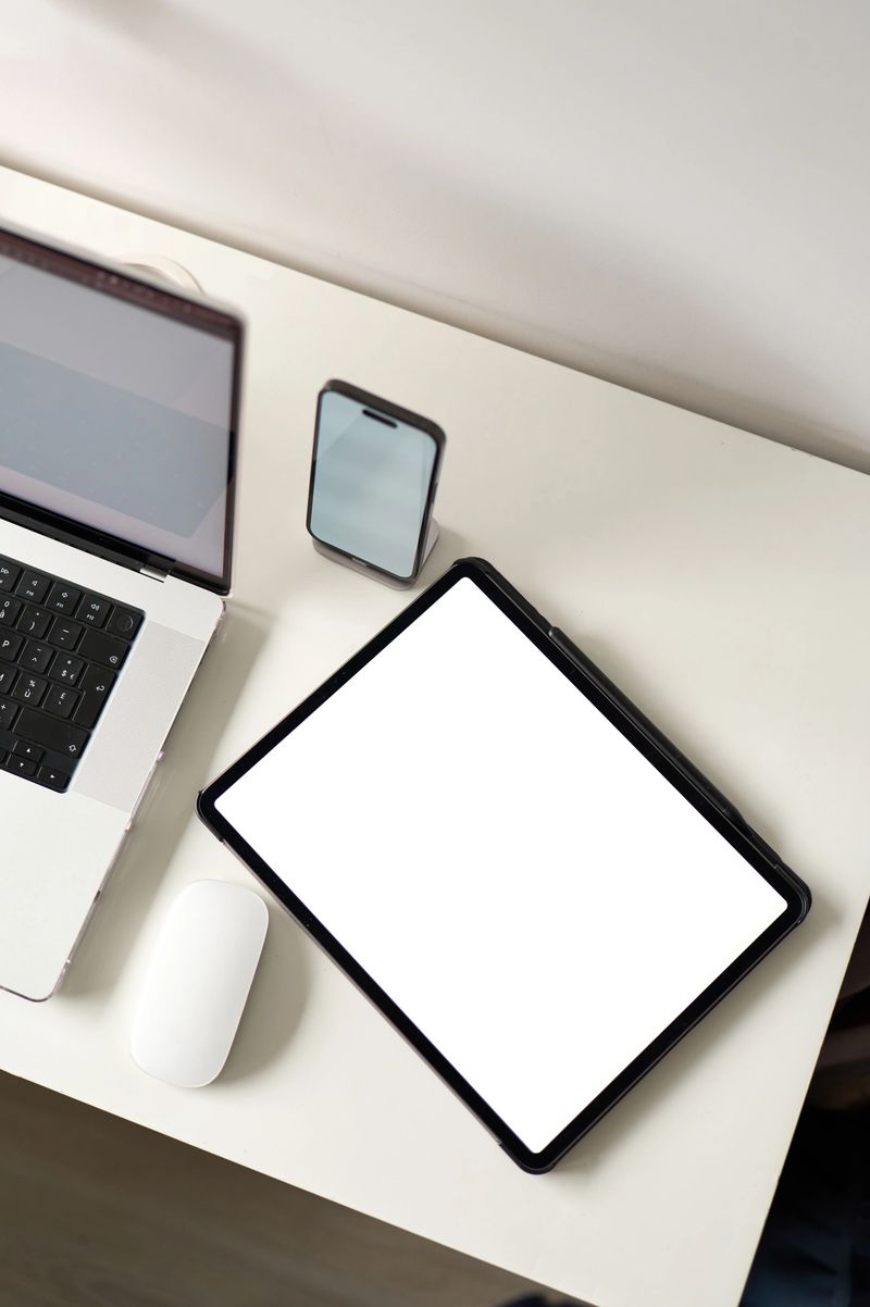 Digital tablet with blank screen, smartphone, laptop and mouse lying on white desk