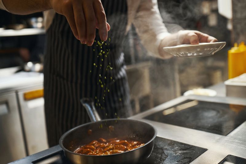 Caucasian middle aged man preparing meal in kitchen, sprinkling chopped herbs into steaming pan of food, wearing apron, holding small plate in one hand, cooking professionally