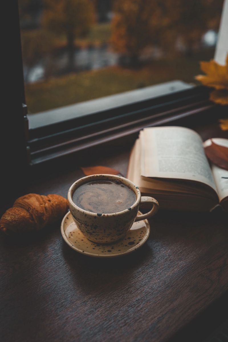 Cozy autumn scene with a ceramic cup of coffee, a croissant, and an open book on a windowsill, with golden leaves and blurred city buildings in the background.