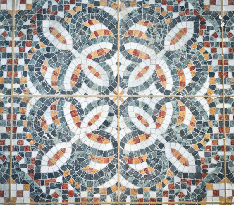 A detailed close-up of a traditional Greek mosaic tile, featuring interlocking geometric loops in earthy tones of green, terracotta, black, and white. This intricate design reflects the harmony and precision of ancient Mediterranean craftsmanship, blending art and symmetry in a timeless floor composition.