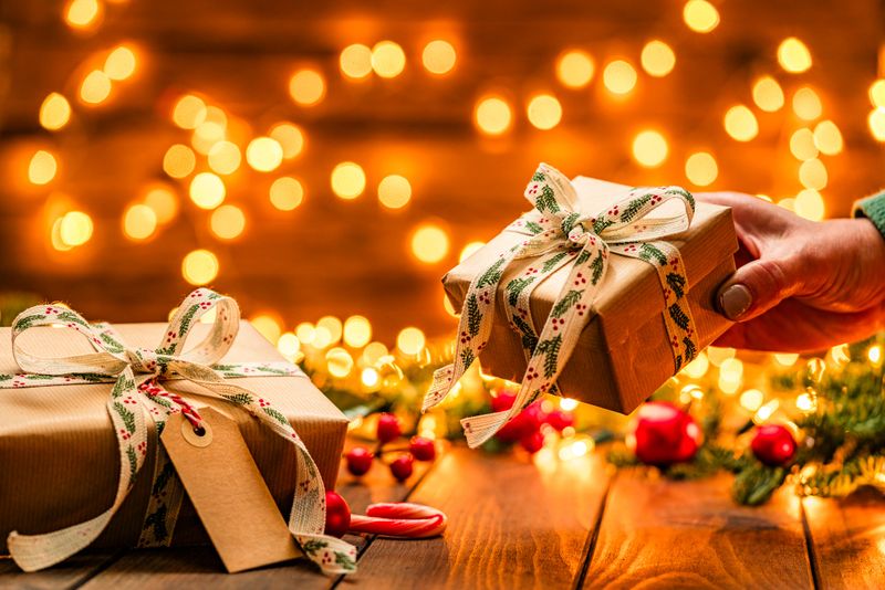 Female hand picking up or giving brown Christmas gift box wrapped with craft paper and white ribbon in a cozy festive atmosphere with defocused Christmas lights at background.