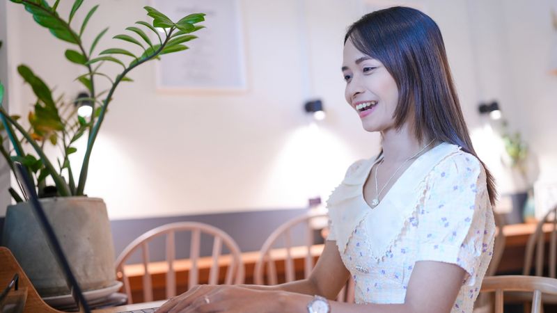 A young woman is smiling while working on laptop in cozy cafe setting. atmosphere is warm and inviting, with plants and soft lighting enhancing ambiance