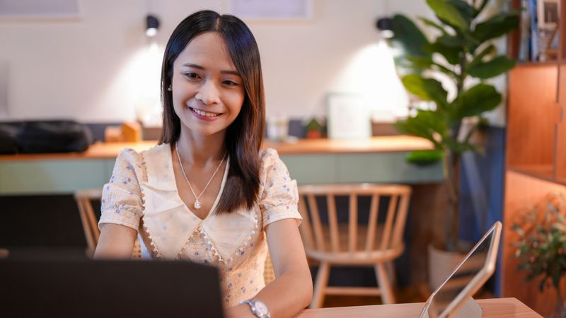 A young woman is smiling while working on laptop in cozy cafe setting. warm lighting and greenery create relaxed atmosphere, perfect for productivity