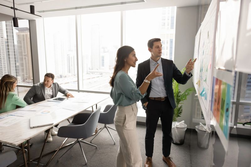 Focused busy Latin business colleagues speaking at Kanban board with sticky notes, discussing tasking, project management, planning teamwork, working on strategy together in meeting room