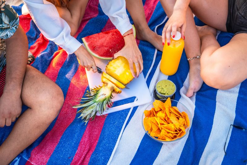 Group of friends having a picnic on a striped towel, cutting pineapple, with watermelon, nachos, guacamole and juice