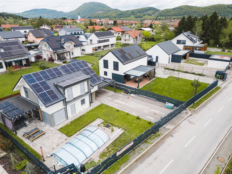Aerial view of a modern residential neighborhood featuring houses with solar panels, green lawns, and well-maintained gardens. The area is surrounded by mountains and has a clear road.