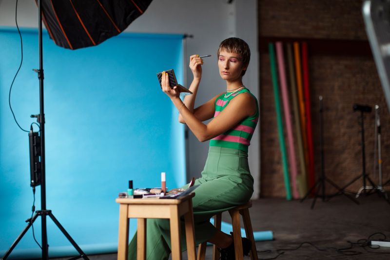 A young gen z girl with short hair applies makeup while sitting in a vibrant photography studio; She wears colorful modern attire, surrounded by professional studio equipment and a blue backdrop