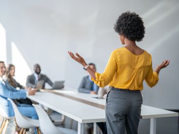 Woman in yellow blouse speaking to business meeting attendees.