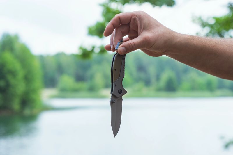 Close-up of man holding folding knife outdoors. Tactical gear and survival tool on a a blurred natural background of trees and water