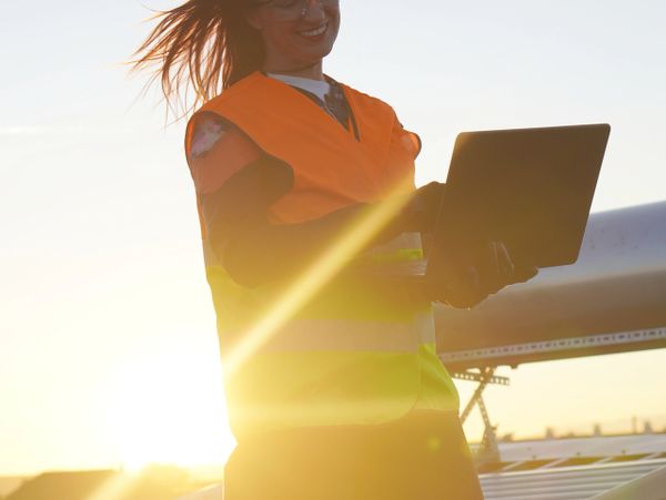 Smiling worker in safety vest using laptop outdoors with sun flare.