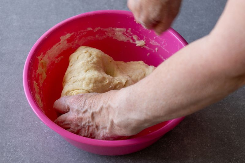 Close-up of an older woman’s hands kneading soft dough in a mixing bowl. Homemade bread or pastry preparation in a warm kitchen setting. Natural light highlights the tactile process of baking.