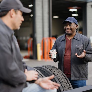 Two mechanics chatting and smiling in a garage, holding coffee cups.