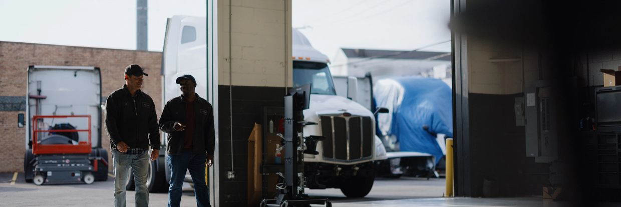 Two men walk and talk inside a truck maintenance garage.