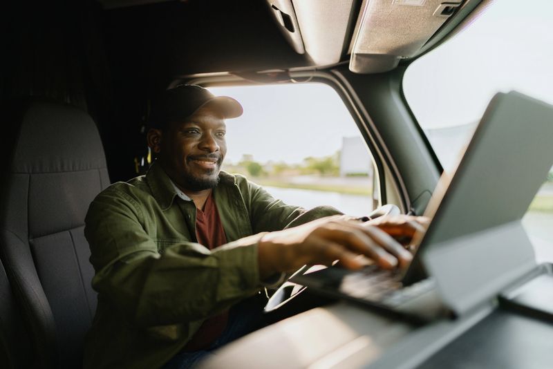 Smiling truck driver using a tablet computer while navigating a semi-truck on a bright, sunny day, enjoying the open road