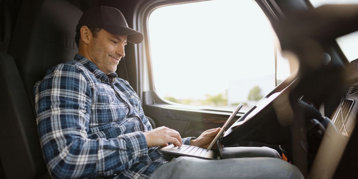Man in a cap using a laptop inside a vehicle, smiling.