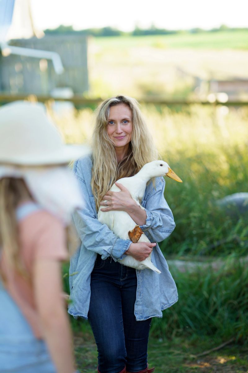 A serene countryside scene featuring a woman holding a white duck, evoking themes of nature, agriculture, and harmony. The green surroundings and a blurred background add an inviting and peaceful ambiance.