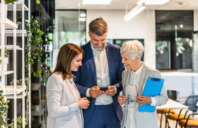 Three professionals smiling and discussing while looking at a phone in a modern office.