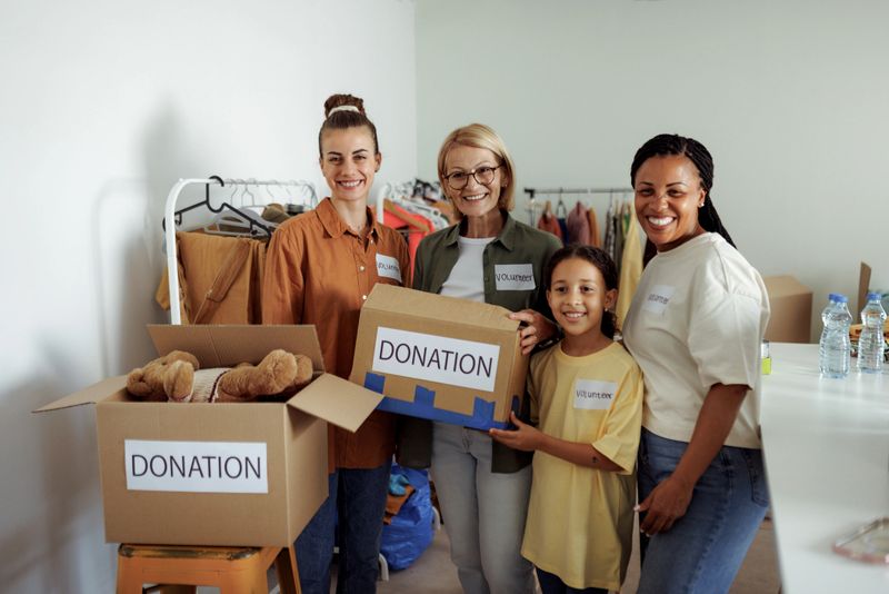 Four female volunteers, including a smiling young girl, posing for the camera in a room filled with donation boxes and a clothes rack. The scene captures teamwork, generosity, and community spirit. Ideal for themes of charity, volunteering, social work, and intergenerational support.
